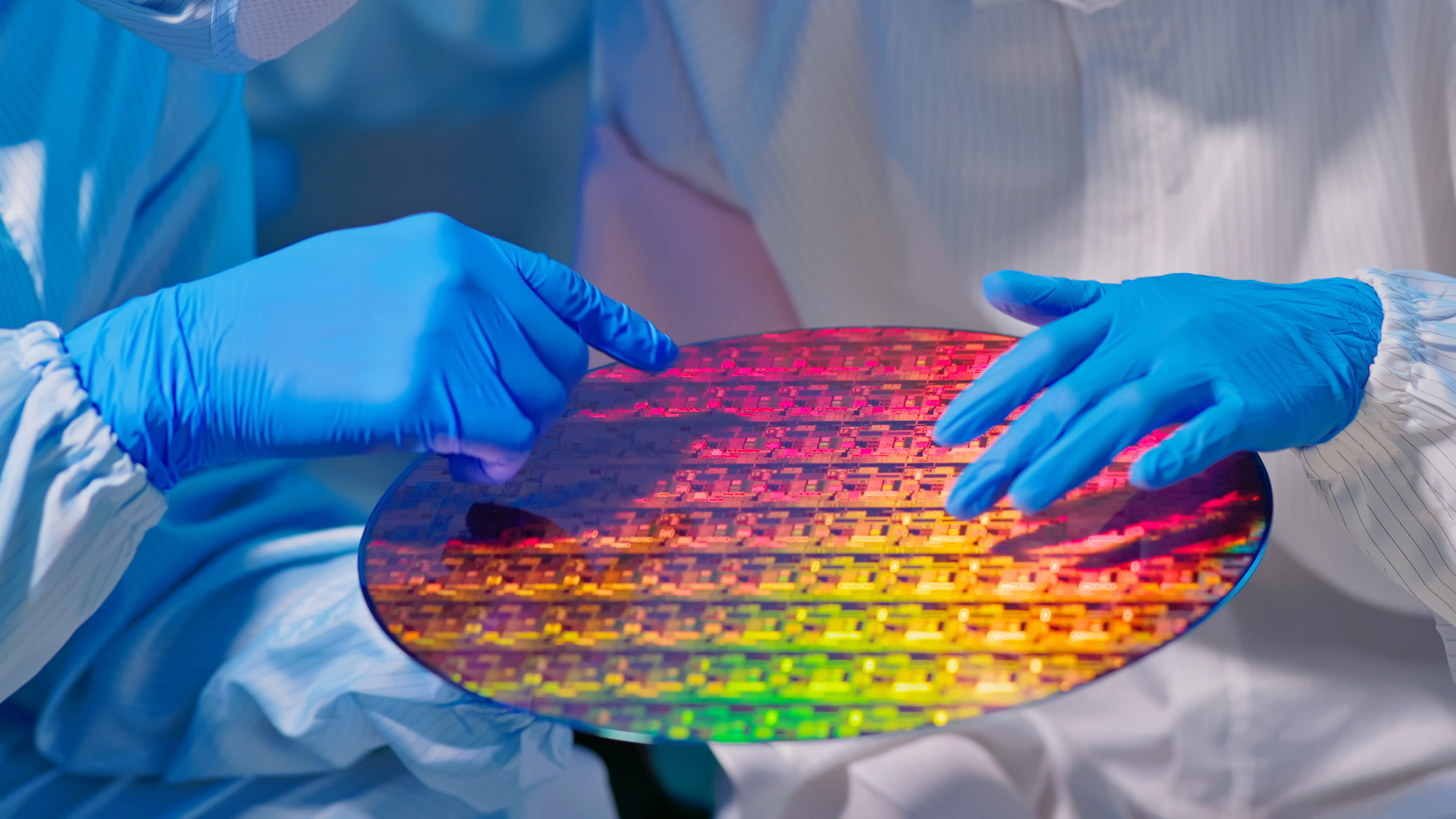 Checking a wafer at a semiconductor manufacturing plant, two technicians in sterile coveralls and gloves hold the wafer, which reflects many different colors.